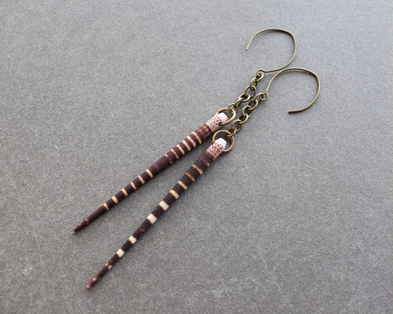 May include: A pair of earrings with brown and white striped, long, thin, teardrop-shaped pendants. The earrings are made with a bronze-colored metal hook.