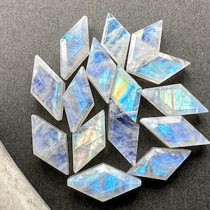 May include: A collection of thirteen translucent, diamond-shaped moonstone cabochons arranged in a cluster. Each gemstone features a faceted surface that displays a distinct iridescent blue and yellow adularescence, reflecting light across the stones.
