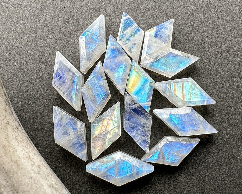 May include: A collection of thirteen translucent, diamond-shaped moonstone cabochons arranged in a cluster. Each gemstone features a faceted surface that displays a distinct iridescent blue and yellow adularescence, reflecting light across the stones.