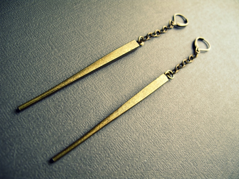 May include: Two long, gold, pointed earrings with chain links. The earrings are laid flat on a gray textured surface.