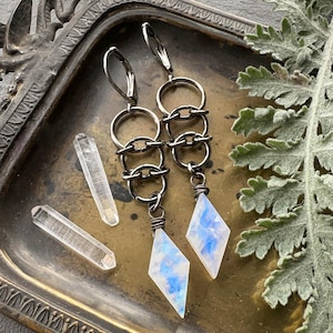 May include: A pair of dangle earrings featuring dark metal lever-back hooks, a series of three interlocking circular rings, and a diamond-shaped moonstone pendant with blue iridescent flashes, accompanied by two small clear quartz crystals.