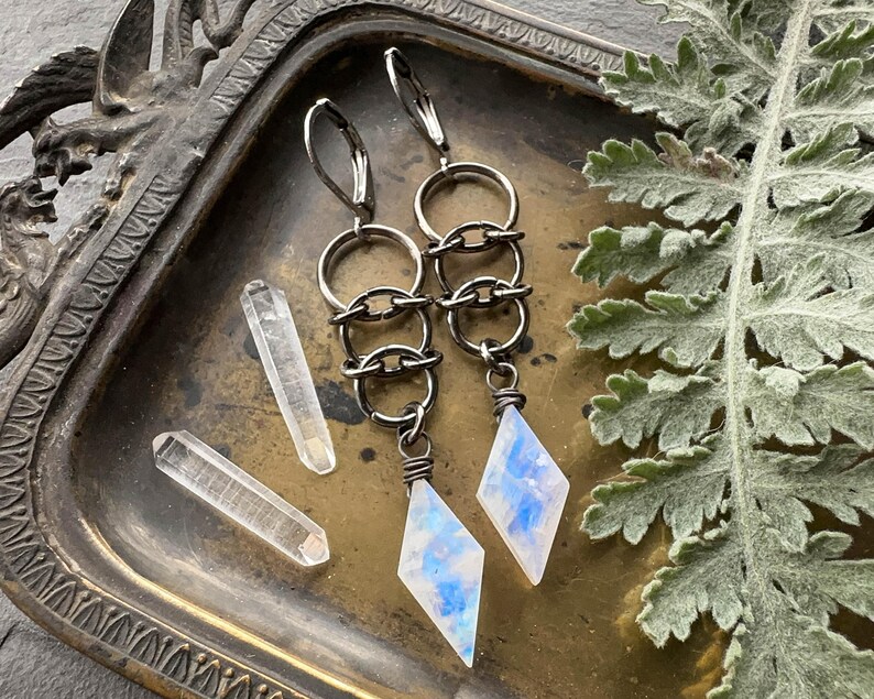 May include: A pair of dangle earrings featuring dark metal lever-back hooks, a series of three interlocking circular rings, and a diamond-shaped moonstone pendant with blue iridescent flashes, accompanied by two small clear quartz crystals.