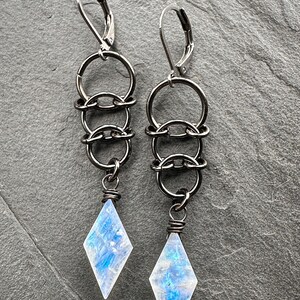 May include: A pair of dangle earrings featuring gunmetal-toned leverback hooks connected to a vertical chain of three interlocking circular rings. Each earring terminates in a faceted, diamond-shaped moonstone pendant with blue iridescence.