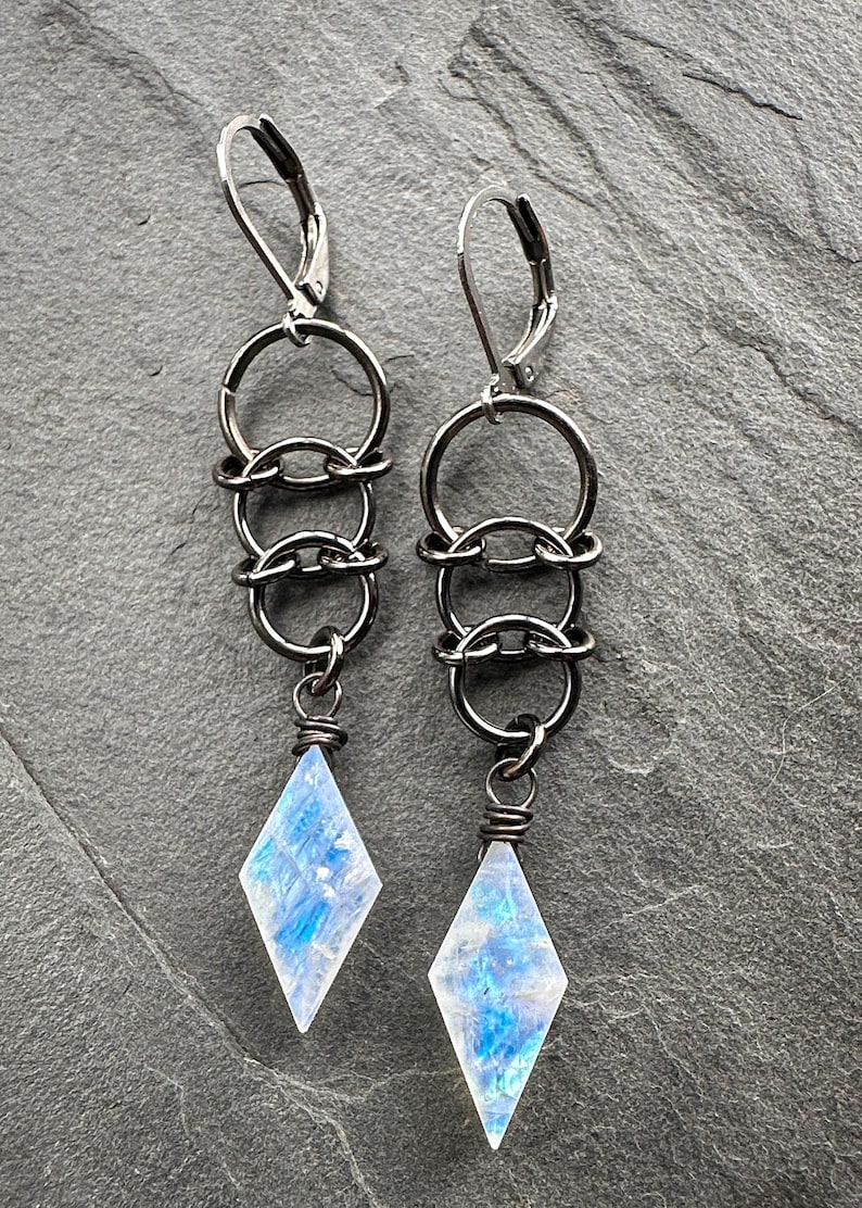 May include: A pair of dangle earrings featuring gunmetal-toned leverback hooks connected to a vertical chain of three interlocking circular rings. Each earring terminates in a faceted, diamond-shaped moonstone pendant with blue iridescence.