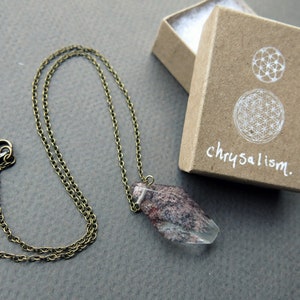 May include: A delicate gold chain necklace with a clear quartz crystal pendant. The crystal has a pink and brown pattern inside. The necklace comes in a small brown box with the word "chrysalism" written on it.
