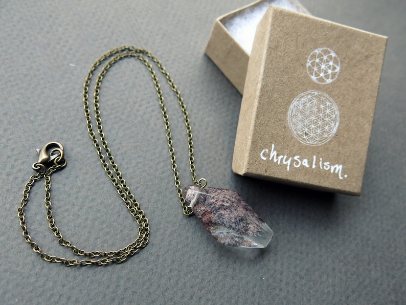 May include: A delicate gold chain necklace with a clear quartz crystal pendant. The crystal has a pink and brown pattern inside. The necklace comes in a small brown box with the word "chrysalism" written on it.