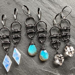 May include: Three pairs of dangle earrings featuring dark gunmetal-toned leverback hooks and interconnected circular metal rings. The first pair has diamond-shaped moonstone drops, the second has teardrop-shaped labradorite, and the third has clear faceted crystal pendants.