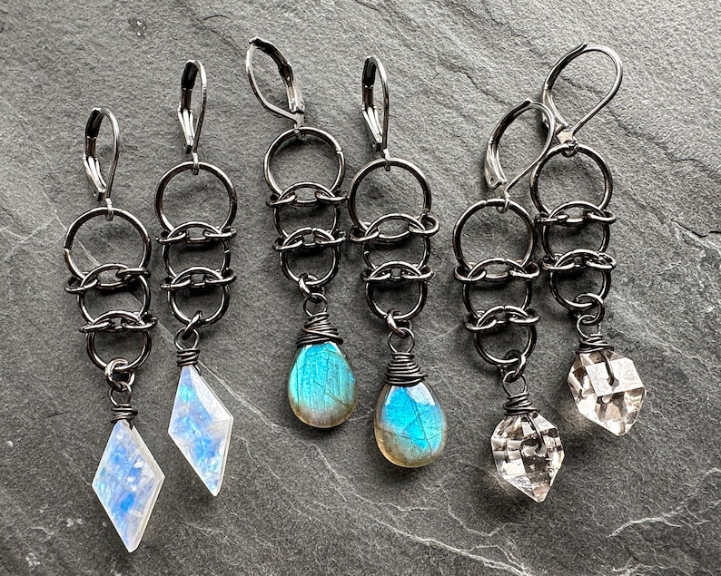 May include: Three pairs of dangle earrings featuring dark gunmetal-toned leverback hooks and interconnected circular metal rings. The first pair has diamond-shaped moonstone drops, the second has teardrop-shaped labradorite, and the third has clear faceted crystal pendants.