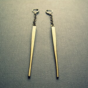 Pu&ograve; includere: Un paio di orecchini pendenti lunghi e sottili, color ottone antico. Ogni orecchino presenta una sottile punta metallica sospesa a una catenella corta e delicata, fissata con una chiusura a monachella, creando un design verticale, elegante e pendente.
