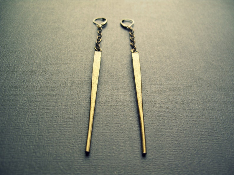 Pu&ograve; includere: Un paio di orecchini pendenti lunghi e sottili, color ottone antico. Ogni orecchino presenta una sottile punta metallica sospesa a una catenella corta e delicata, fissata con una chiusura a monachella, creando un design verticale, elegante e pendente.
