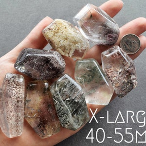 May include: A collection of eight clear quartz crystals with inclusions, ranging in size from 40 to 55 millimeters. The crystals are in various shapes and have a variety of inclusions, including black, brown, and green. The crystals are displayed on a hand.