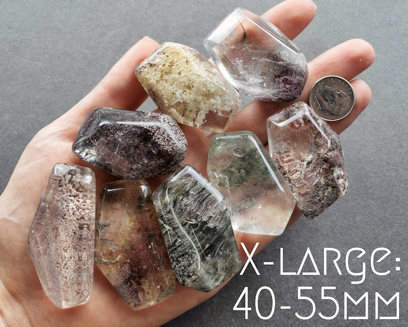 May include: A collection of eight clear quartz crystals with inclusions, ranging in size from 40 to 55 millimeters. The crystals are in various shapes and have a variety of inclusions, including black, brown, and green. The crystals are displayed on a hand.