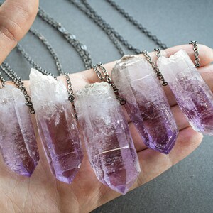 May include: Six amethyst crystal pendants on silver chains. The crystals are pointed and have a light purple color.
