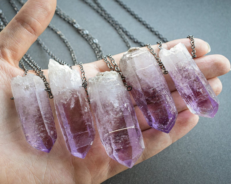 May include: Six amethyst crystal pendants on silver chains. The crystals are pointed and have a light purple color.