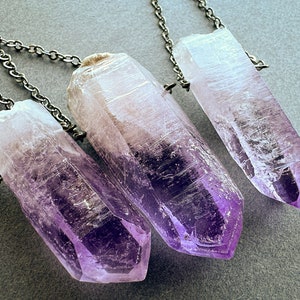 May include: Three amethyst crystal points on silver chains. The crystals are clear with a purple hue and have a pointed tip. The chains are delicate and have a silver finish.