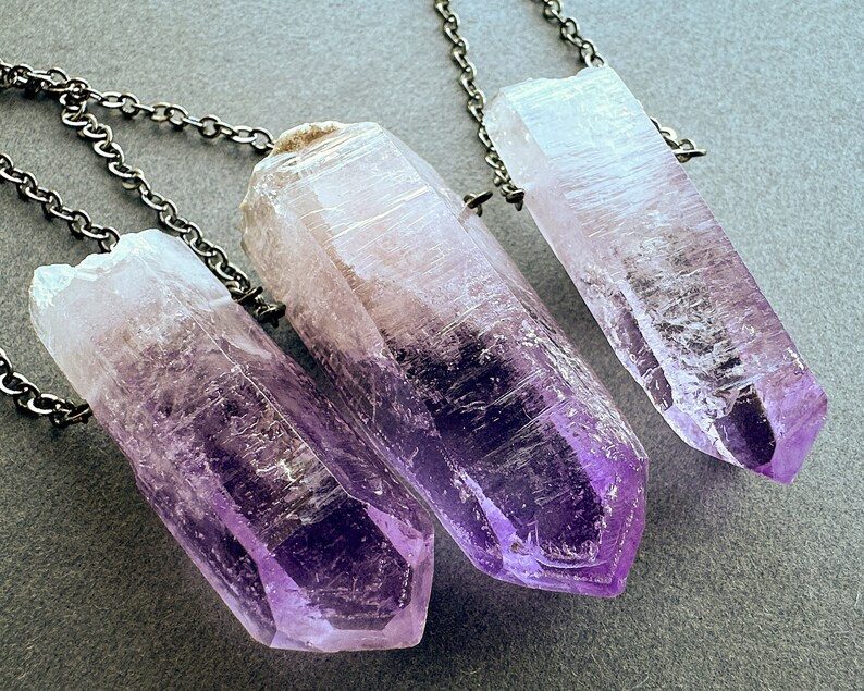 May include: Three amethyst crystal points on silver chains. The crystals are clear with a purple hue and have a pointed tip. The chains are delicate and have a silver finish.