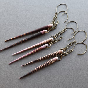 May include: Three pairs of earrings with long, thin, striped pendants. The pendants are made of natural materials and have a brown and pink color scheme. The earrings are attached to gold-toned chains with hook closures.
