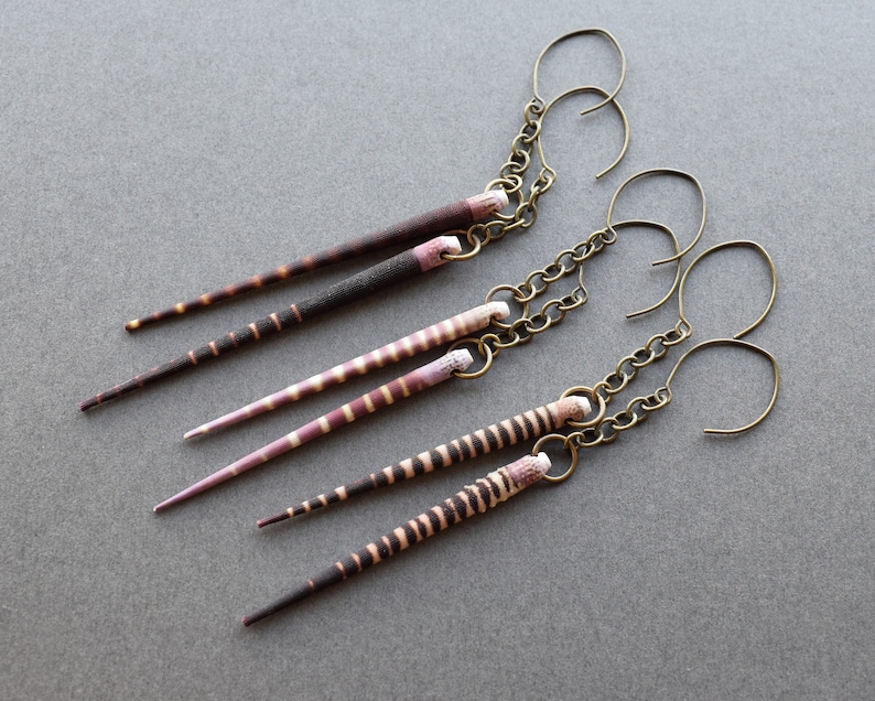 May include: Three pairs of earrings with long, thin, striped pendants. The pendants are made of natural materials and have a brown and pink color scheme. The earrings are attached to gold-toned chains with hook closures.