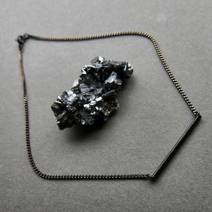 May include: A black chain necklace with a black bar pendant. The necklace is on a gray surface with a black crystal.