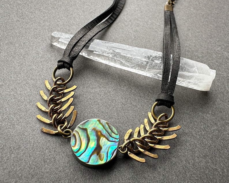 May include: A necklace with a black leather cord and a large iridescent abalone shell pendant. The pendant is surrounded by a brass chain with decorative spikes.