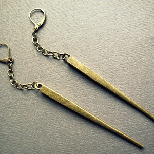 Pu&ograve; includere: Un paio di orecchini pendenti con lunghe barre metalliche sottili e affusolate in finitura ottone anticato. Ogni barra &egrave; sospesa a una corta e delicata catena a maglie collegata a una chiusura a monachella, creando un design lineare.