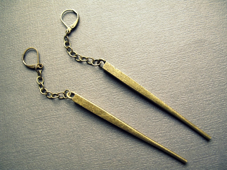 Pu&ograve; includere: Un paio di orecchini pendenti con lunghe barre metalliche sottili e affusolate in finitura ottone anticato. Ogni barra &egrave; sospesa a una corta e delicata catena a maglie collegata a una chiusura a monachella, creando un design lineare.