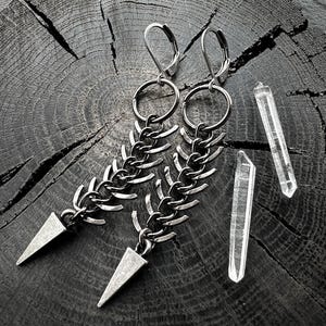 May include: Silver-toned earrings with a chain design and triangular pendants. The earrings are displayed with two clear quartz crystal points on a dark, textured wooden surface. The earrings have a lever back closure.