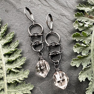 May include: A pair of dangle earrings featuring dark gunmetal-toned leverback hooks connected to a vertical chain of three interlocking circular rings, finished with a single clear, faceted, diamond-shaped crystal drop at the base.