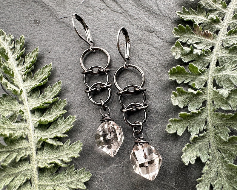 May include: A pair of dangle earrings featuring dark gunmetal-toned leverback hooks connected to a vertical chain of three interlocking circular rings, finished with a single clear, faceted, diamond-shaped crystal drop at the base.