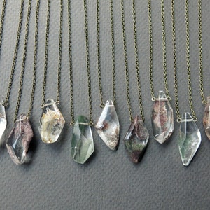 May include: Ten necklaces with clear quartz crystal pendants featuring inclusions of green, brown, and white. The pendants are faceted and have a geometric shape. The chains are silver-toned.