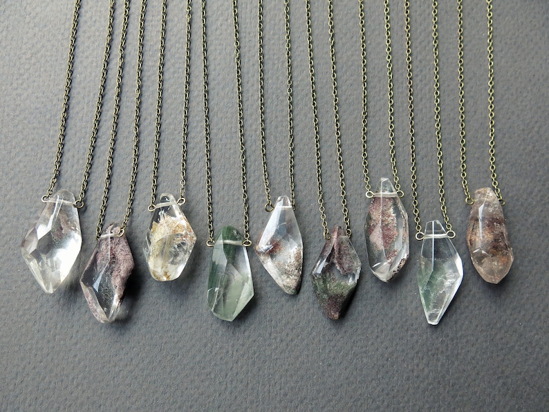 May include: Ten necklaces with clear quartz crystal pendants featuring inclusions of green, brown, and white. The pendants are faceted and have a geometric shape. The chains are silver-toned.