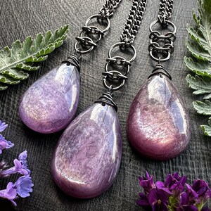 K&ouml;nnte beinhalten: Drei tropfenf&ouml;rmige Anh&auml;nger in schimmerndem Lila. Jeder Anh&auml;nger h&auml;ngt an einer Kette aus Gunmetal mit einem dekorativen Glied. Die Anh&auml;nger sind oben mit Draht umwickelt. Das Bild ist mit lila Blumen und gr&uuml;nem Laub gestaltet.
