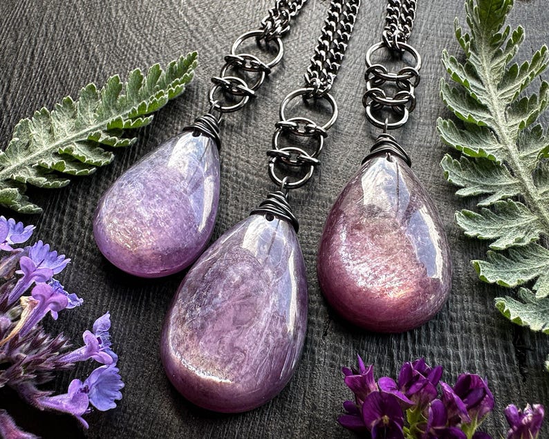 K&ouml;nnte beinhalten: Drei tropfenf&ouml;rmige Anh&auml;nger in schimmerndem Lila. Jeder Anh&auml;nger h&auml;ngt an einer Kette aus Gunmetal mit einem dekorativen Glied. Die Anh&auml;nger sind oben mit Draht umwickelt. Das Bild ist mit lila Blumen und gr&uuml;nem Laub gestaltet.