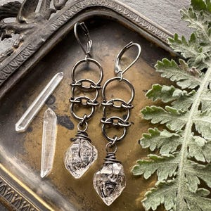 May include: A pair of dark metal dangle earrings featuring a series of three interlocking circular rings connected to a raw, clear quartz crystal point wrapped in dark wire at the base, with lever-back closures for secure attachment.