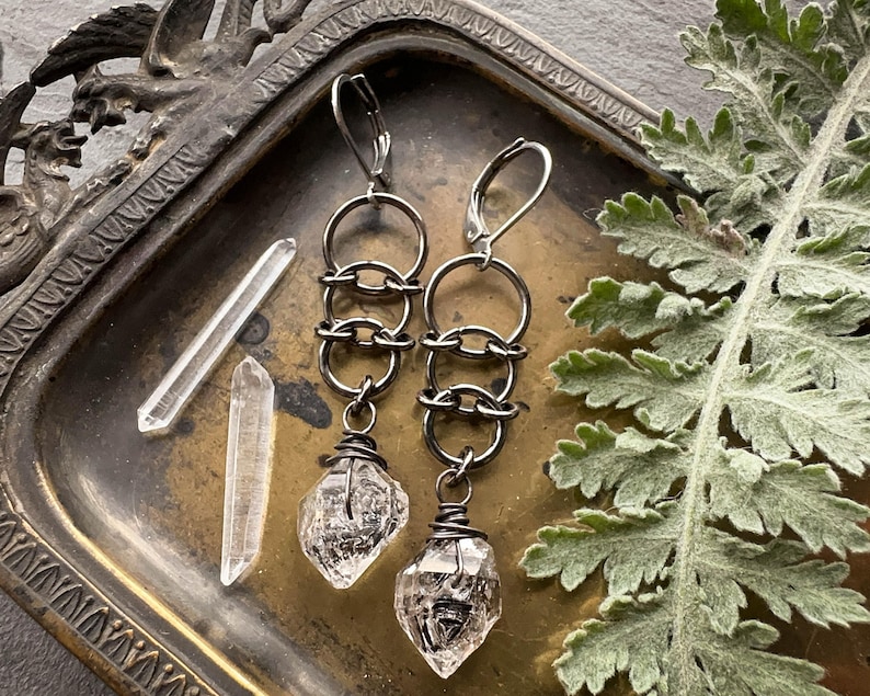 May include: A pair of dark metal dangle earrings featuring a series of three interlocking circular rings connected to a raw, clear quartz crystal point wrapped in dark wire at the base, with lever-back closures for secure attachment.