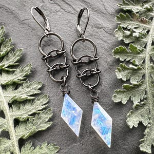 May include: A pair of dangle earrings featuring dark gunmetal-toned leverback hooks connected to three interlocking circular rings, ending in a faceted, diamond-shaped moonstone pendant with iridescent blue and white flashes.