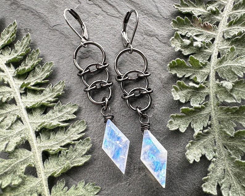 May include: A pair of dangle earrings featuring dark gunmetal-toned leverback hooks connected to three interlocking circular rings, ending in a faceted, diamond-shaped moonstone pendant with iridescent blue and white flashes.