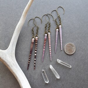 May include: Four pairs of dangle earrings with a long, thin, striped shell pendant. The shells are in shades of pink and brown. The earrings are hanging from a silver chain and hook.