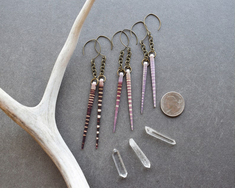 May include: Four pairs of dangle earrings with a long, thin, striped shell pendant. The shells are in shades of pink and brown. The earrings are hanging from a silver chain and hook.