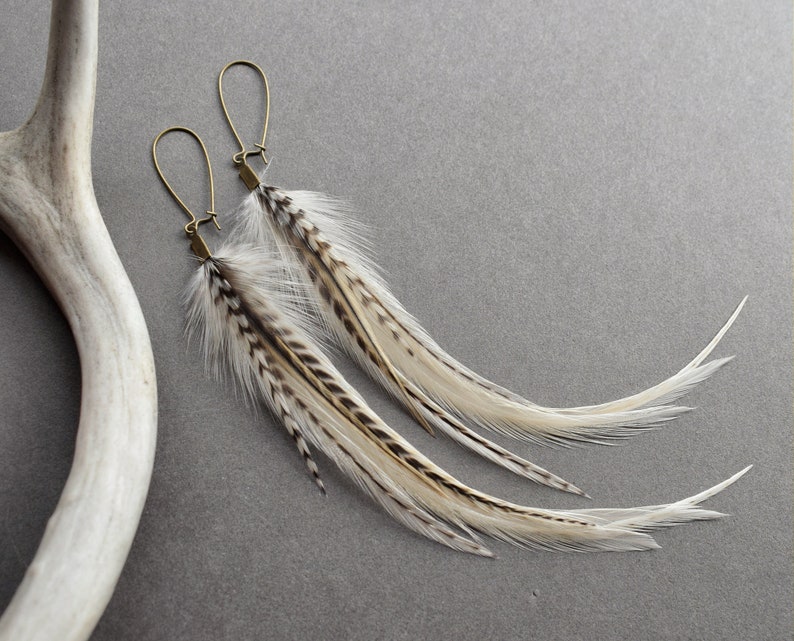 May include: A pair of long, white and brown feather earrings with gold-toned hoops. The feathers are arranged in a cascading pattern.