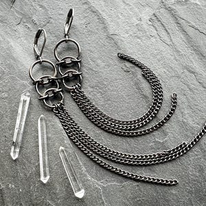 May include: A pair of silver-toned dangle earrings with multiple chain tassels. The earrings feature three connected circular hoops and are accompanied by three clear quartz crystal points. The jewelry is displayed on a gray stone surface.