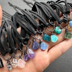 K&ouml;nnte beinhalten: Eine Sammlung von Lederkordelketten mit verschiedenen Edelsteinanh&auml;ngern, darunter Amethyst, T&uuml;rkis, Aquamarin und Labradorit. Die Ketten werden auf einer Hand pr&auml;sentiert.
