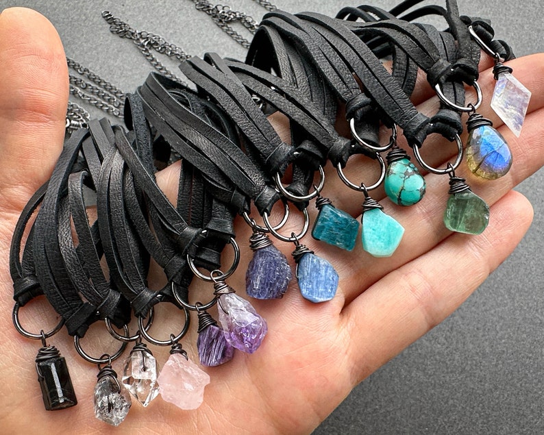 K&ouml;nnte beinhalten: Eine Sammlung von Lederkordelketten mit verschiedenen Edelsteinanh&auml;ngern, darunter Amethyst, T&uuml;rkis, Aquamarin und Labradorit. Die Ketten werden auf einer Hand pr&auml;sentiert.