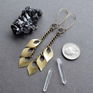 May include: A pair of long, gold-toned earrings with a chain and multiple leaf-shaped charms. The earrings are dangling and have a simple, elegant design.