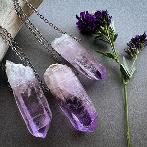 May include: Three amethyst crystal point necklaces with silver chains. The crystals are various shades of purple and are hanging from a white surface.