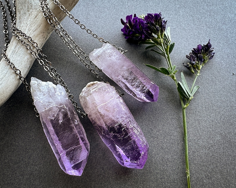 May include: Three amethyst crystal point necklaces with silver chains. The crystals are various shades of purple and are hanging from a white surface.