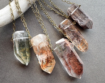 May include: Close-up of several crystal pendant necklaces. Each necklace features a raw, clear quartz crystal pendant suspended from a gold-coloured chain. The crystals vary in colour, with some showing green, brown, and reddish hues.