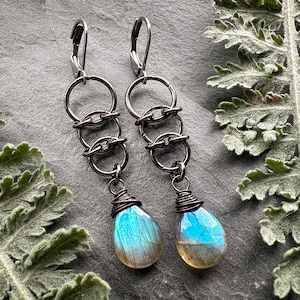 May include: A pair of drop earrings featuring dark gunmetal-toned leverback hooks connected to three interlocking circular rings. Each earring terminates in a teardrop-shaped labradorite gemstone with a blue iridescent flash, wrapped in wire.