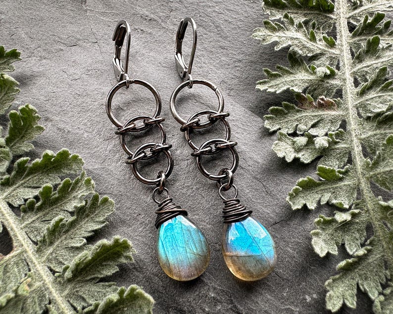 May include: A pair of drop earrings featuring dark gunmetal-toned leverback hooks connected to three interlocking circular rings. Each earring terminates in a teardrop-shaped labradorite gemstone with a blue iridescent flash, wrapped in wire.