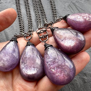 K&ouml;nnte beinhalten: Sechs tropfenf&ouml;rmige Amethyst-Anh&auml;nger an Silberketten. Jeder Anh&auml;nger hat eine andere Gr&ouml;&szlig;e und eine drahtumwickelte &Ouml;se. Die Amethyststeine haben eine durchscheinende, violette Farbe mit inneren Mustern. Die Ketten sind dunkelsilberfarben.